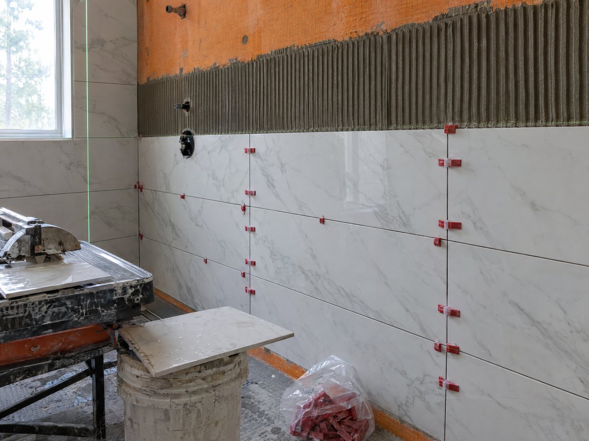 Large-format marble-look porcelain being set on a shower wall over orange membrane, red leveling clips at the joints, wet saw and thinset bucket